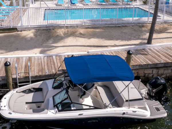 A motorboat with a blue canopy is docked near a wooden boardwalk, with a fenced pool area and lounge chairs in the background.