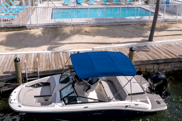 A motorboat with a blue canopy is docked near a wooden boardwalk, with a fenced pool area and lounge chairs in the background.