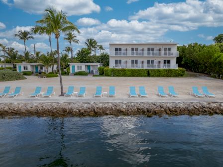 A beachfront resort with palm trees, lounge chairs, and a multi-story building facing the water. The sky is partly cloudy and the water is calm.