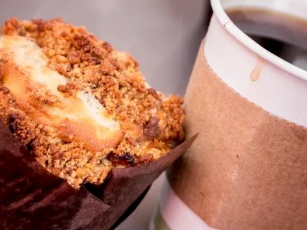 The image shows a muffin with a crumbly topping next to a cup of coffee in a paper cup with a cardboard sleeve.