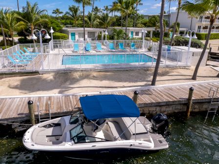 The image shows a boat docked by a waterfront, with a pool enclosed by a white fence, surrounded by lounge chairs and palm trees in the background.