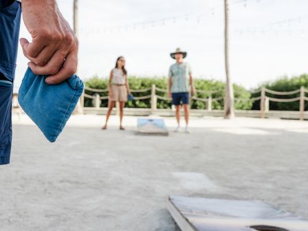 People play a game outdoors, with one holding a bean bag, aiming towards a cornhole board in the foreground.