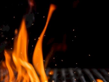 The image shows bright orange flames against a dark background, possibly from a grill, with small sparks visible.