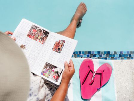 A person is sitting by the pool, reading a magazine. Nearby are pink flip-flops on a striped towel, and their feet are dipped in the water.