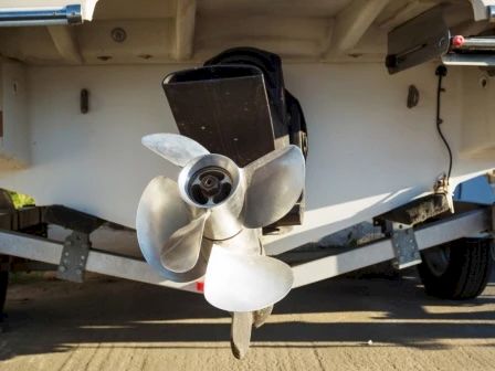 The image shows the back end of a boat with an outboard motor and a three-blade propeller, resting on a trailer out of the water.