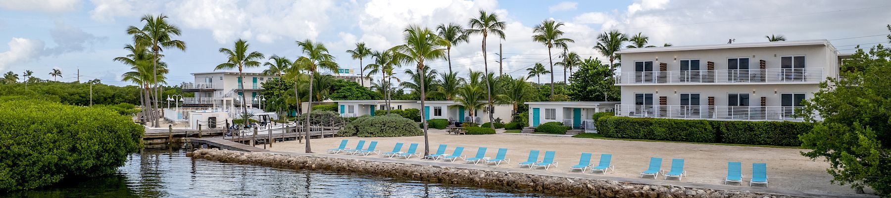 A waterfront view of a serene beachside resort with lounge chairs, palm trees, and two white buildings under a partly cloudy sky, reflected in calm water.