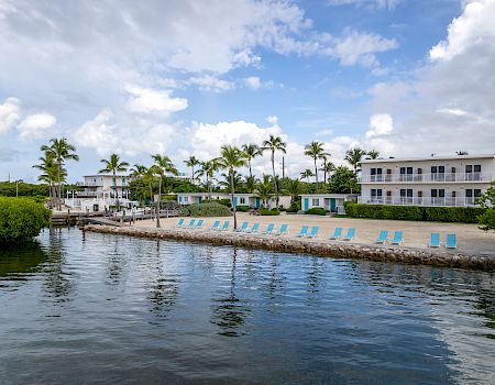 A waterfront view of a serene beachside resort with lounge chairs, palm trees, and two white buildings under a partly cloudy sky, reflected in calm water.