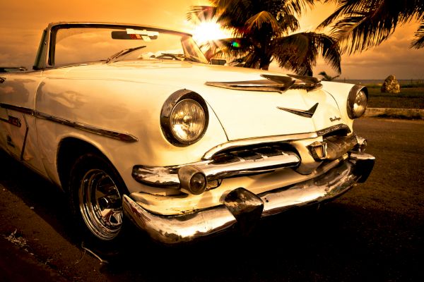 A vintage white convertible car is parked near a beach at sunset, with palm trees and the ocean in the background.