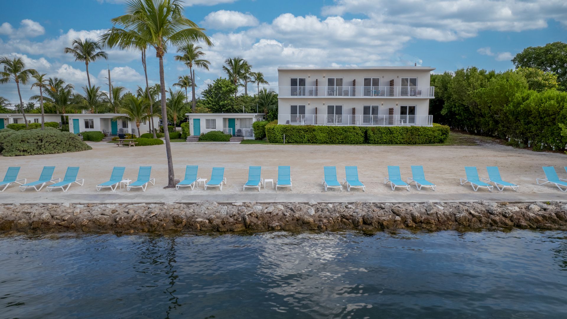 A beachfront property with palm trees, lounge chairs lined up along the shore, and a two-story building with balconies, under a partly cloudy sky.