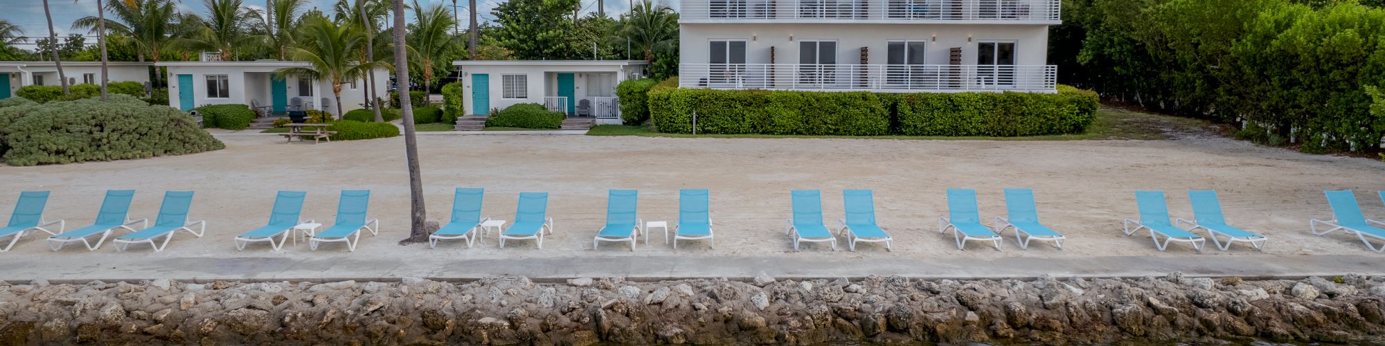 A beachfront property with palm trees, lounge chairs lined up along the shore, and a two-story building with balconies, under a partly cloudy sky.