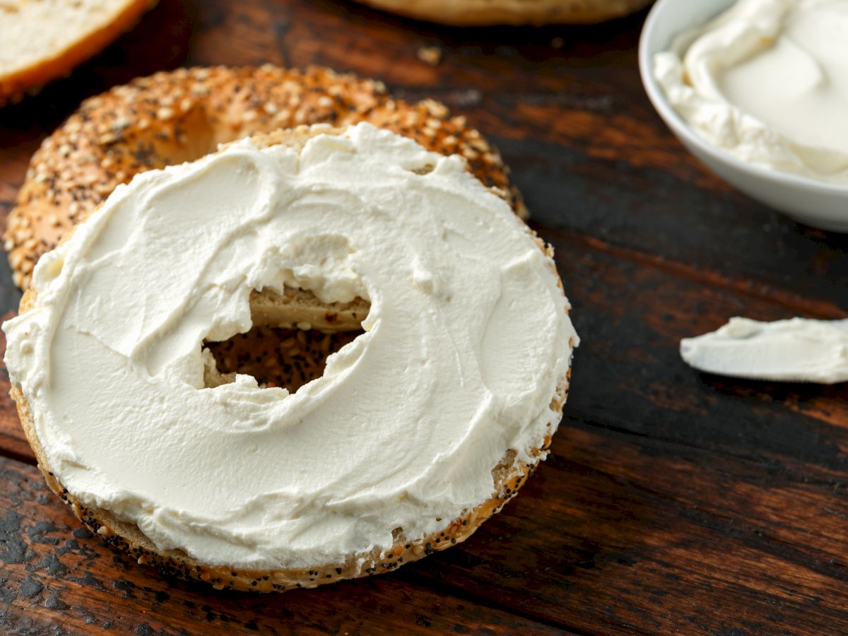 A bagel with cream cheese spread, placed on a wooden surface, next to a knife and a bowl of additional cream cheese.