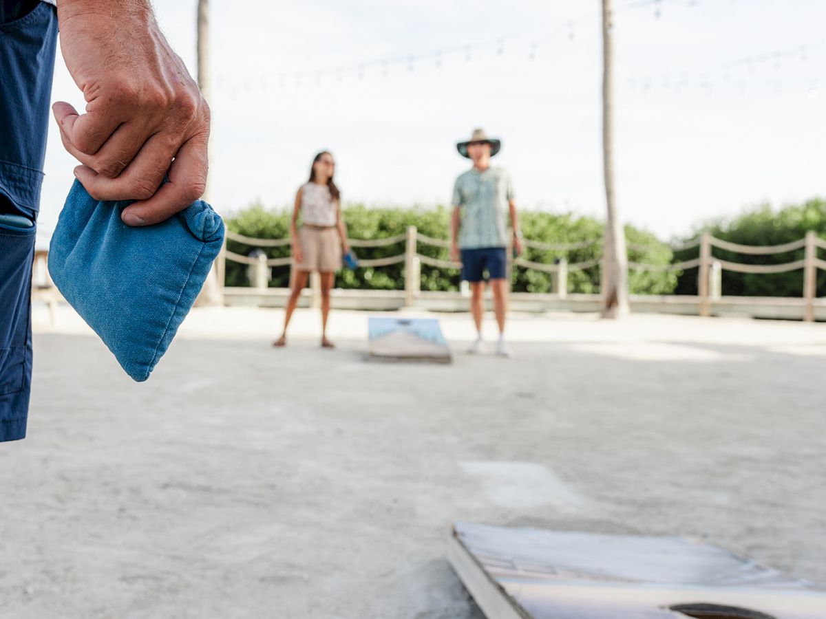 People playing a game of cornhole outdoors, with one person holding a blue bean bag and others in the background near a cornhole board.