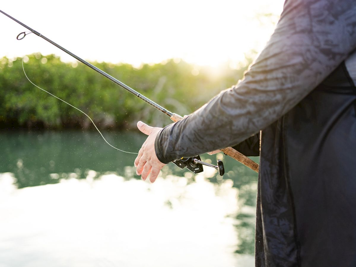 A person is fishing near a water body with a fishing rod in hand, the background shows greenery and sunlight reflecting off the water.