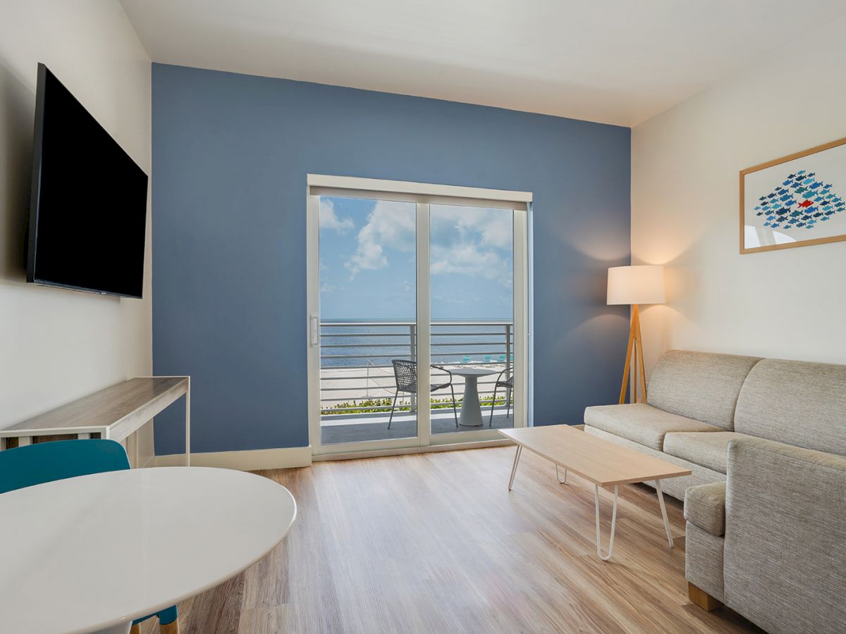 A modern living room with a blue accent wall, TV, sofa, lamp, coffee table, and glass doors leading to a balcony with a beach view.