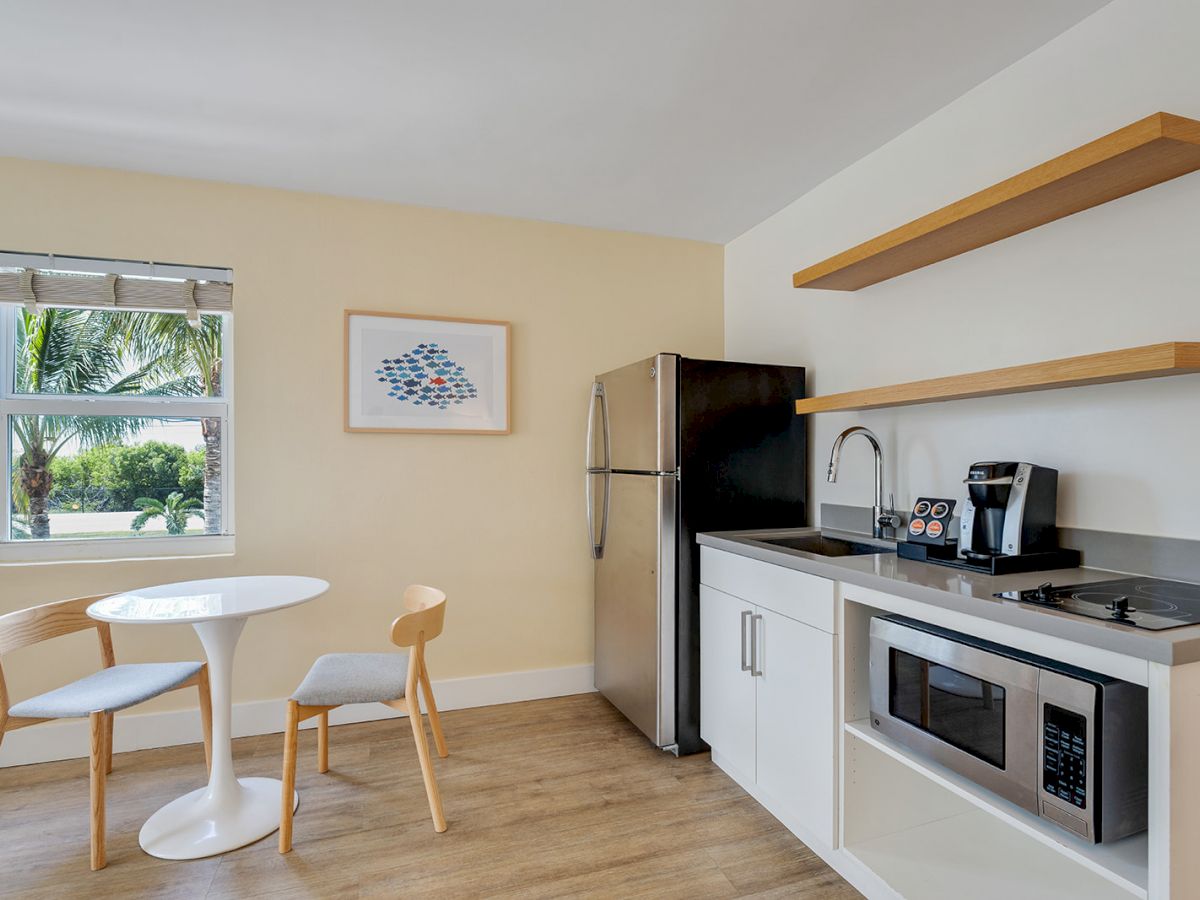 A modern kitchen with a small table and two chairs, a refrigerator, a microwave, open shelves, and a window with a view of palm trees, ending the sentence.