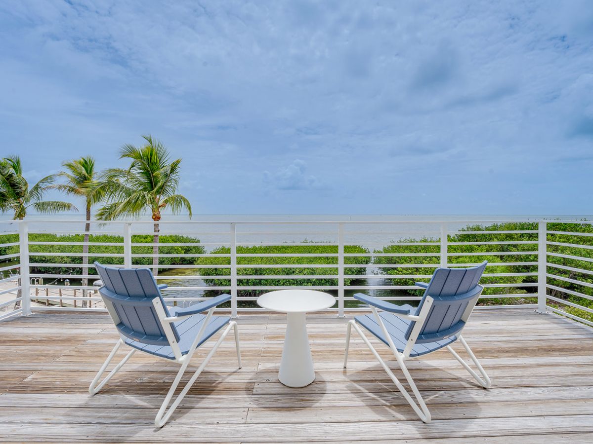 Two chairs and a small table on a wooden deck overlook the ocean, with palm trees and greenery in the background, under a partly cloudy sky.