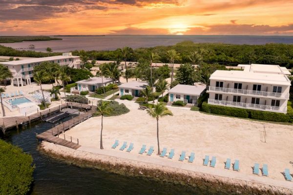 An aerial view of a beachfront resort with buildings, a pool, palm trees, and lounge chairs on the sandy shore during a golden hour sunset.