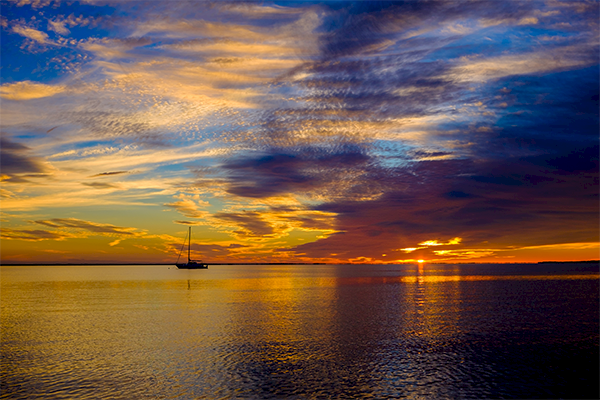 A serene sunset over calm water with a sailboat silhouetted on the horizon, surrounded by vibrant, colorful clouds.