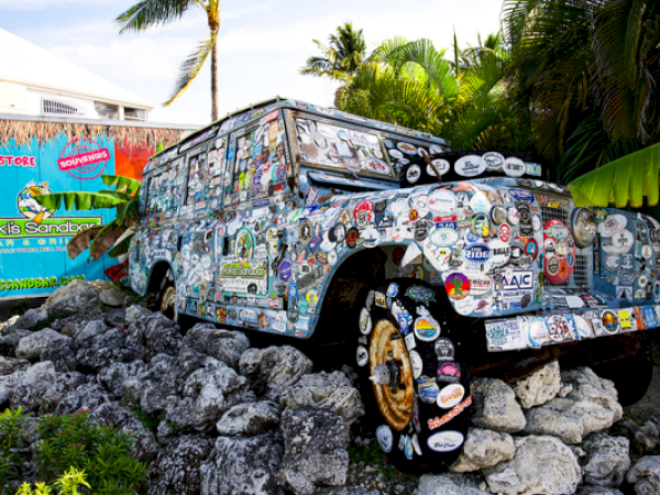 A sticker-covered vehicle is parked on rocks near a colorful sign. It’s surrounded by lush greenery and palm trees.