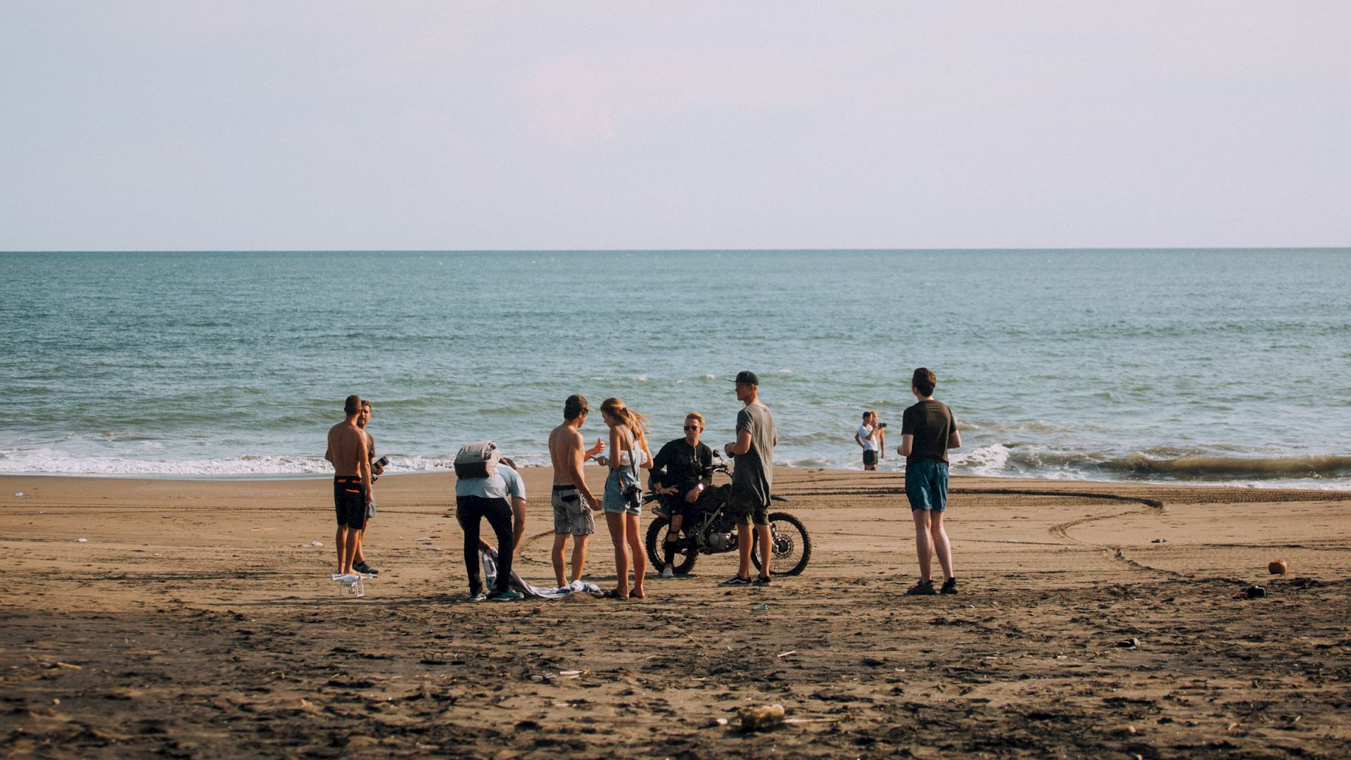 A group of people stands on a sandy beach near the ocean, with a motorcycle and a drone flying above them in the distance.