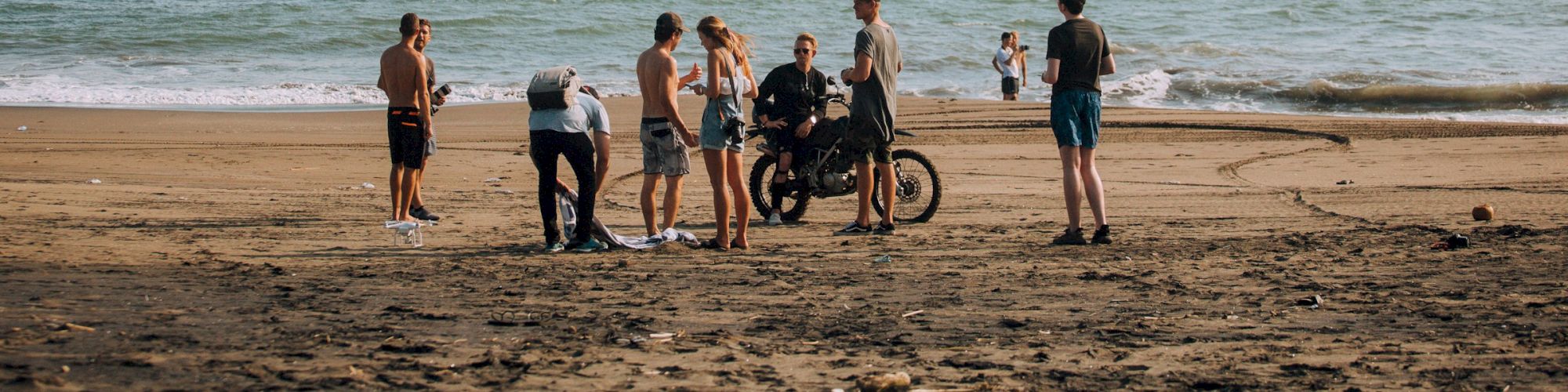 A group of people stands on a sandy beach near the ocean, with a motorcycle and a drone flying above them in the distance.