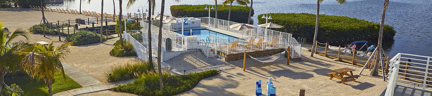 A beachside area with palm trees, a pool, picnic tables, and cornhole games, overlooking a calm body of water.
