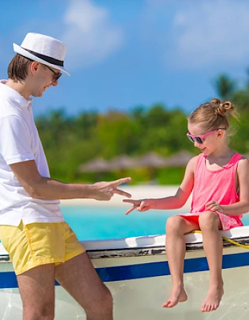 A man and a child are playing rock-paper-scissors on a boat by a beach, surrounded by clear blue water and a tropical setting.