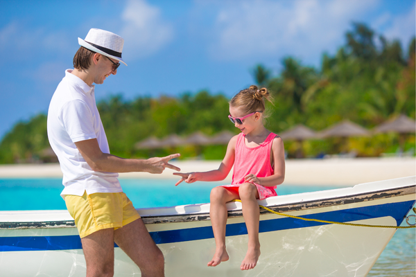 A man and a girl play rock-paper-scissors on a boat by the beach, with clear skies and water in the background.