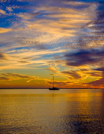 A sailboat on calm waters during a vivid sunset with dramatic clouds and reflections on the sea surface.