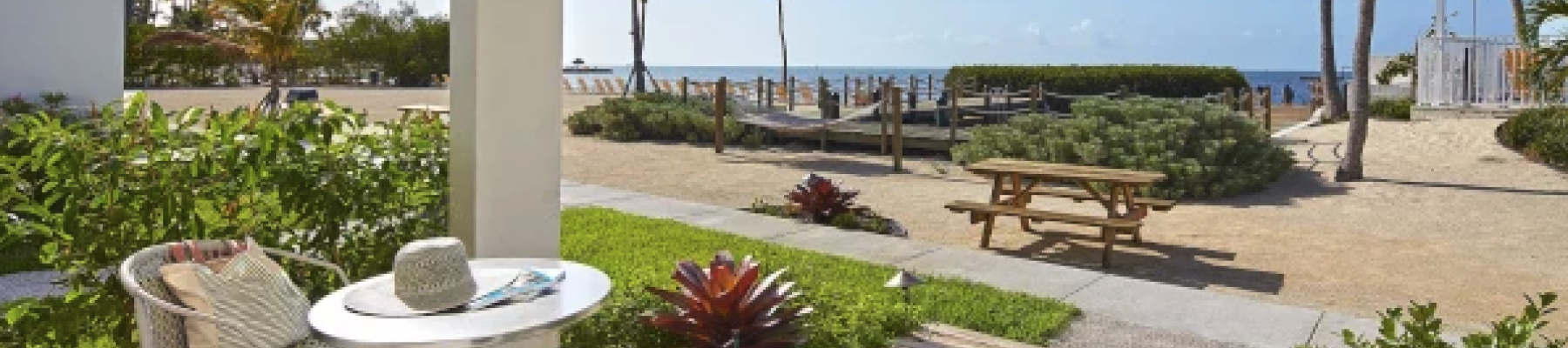 A patio with a table and chair overlooks a pathway leading to the beach, surrounded by tropical plants and palm trees under a blue sky.