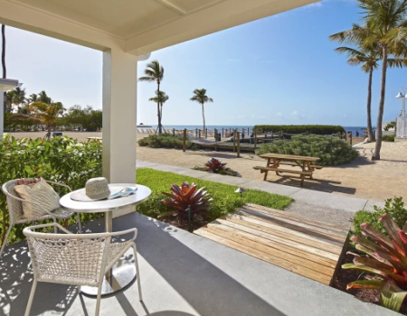 A patio with a table and chair overlooks a pathway leading to the beach, surrounded by tropical plants and palm trees under a blue sky.