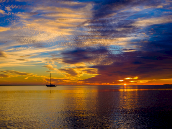 A sailboat on calm water at sunset with vibrant, colorful clouds in the sky.