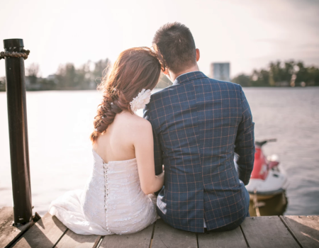 A couple sits on a dock, facing a serene lake, with the sun setting in the background.