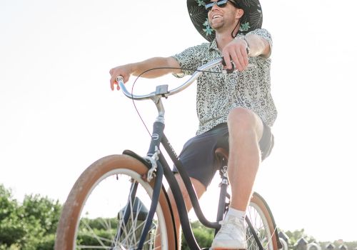 A stylish person rides a vintage bicycle with wide tan tires along a sunny boardwalk, wearing a hat, sunglasses, and patterned shirt, enjoying the breeze.