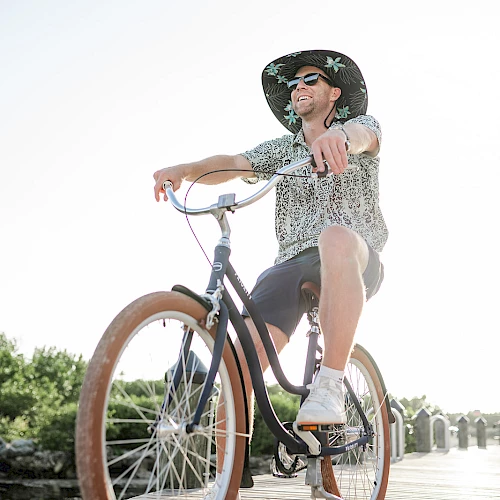 A stylish person rides a vintage bicycle with wide tan tires along a sunny boardwalk, wearing a hat, sunglasses, and patterned shirt, enjoying the breeze.