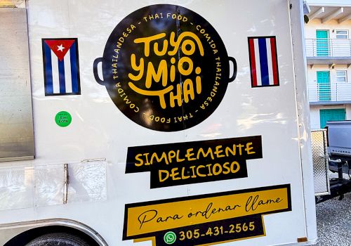 A white food truck with a round black and yellow logo, Cuban flags, and yellow banners advertising &ldquo;TUYO Y MIO, THAI&rdquo; and &ldquo;SIMPLEMENTE DELICIOSO,&rdquo; plus a phone number.