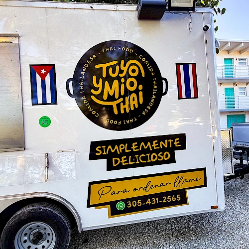 A white food truck with a round black and yellow logo, Cuban flags, and yellow banners advertising &ldquo;TUYO Y MIO, THAI&rdquo; and &ldquo;SIMPLEMENTE DELICIOSO,&rdquo; plus a phone number.