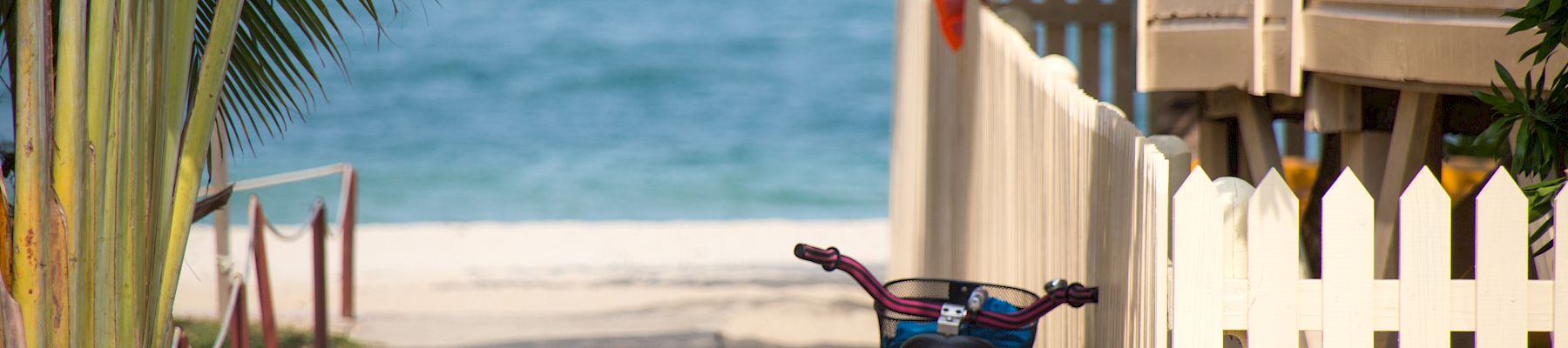 A sandy path leads to the blue sea, framed by palm leaves and a white fence, with a bicycle resting near the entrance to the beach.