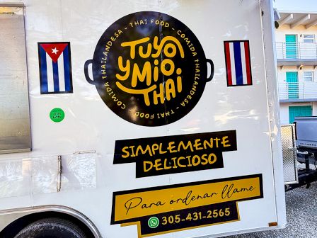 A white food truck with a circular black and gold logo, Cuban flags, yellow banners, and a phone number for &ldquo;Tuyo y M&iacute;o Thai&rdquo; &mdash; simple, delicious.