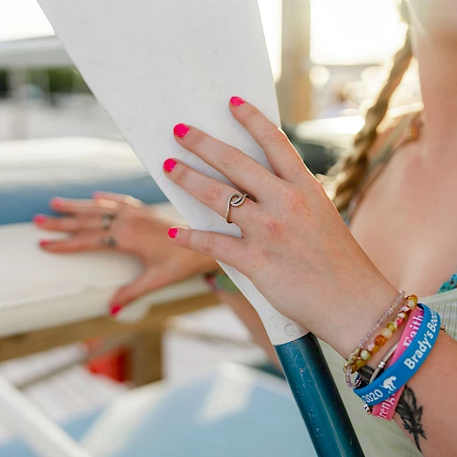 A woman with pink nail polish holds a white paddle by a pool, wearing bracelets and a blue swimsuit, with braided hair and a ring.