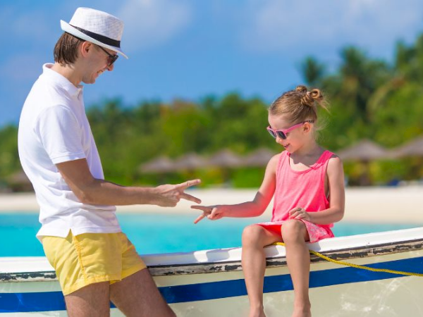 A man in a white shirt and yellow shorts offers a ring to a girl in a pink dress as they share a playful moment on a boat by the beach.