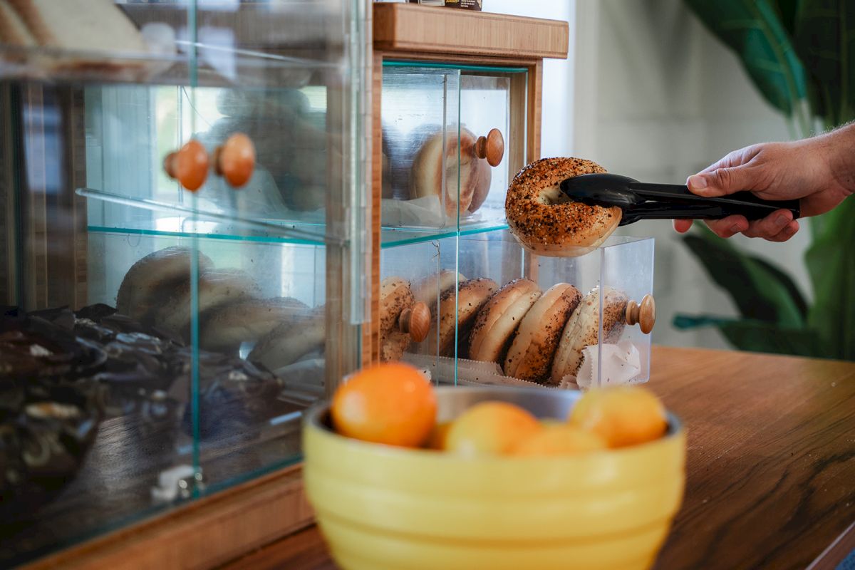 A person uses tongs to grab a bagel from a glass display case at a bakery, with a bowl of oranges and pastries nearby.
