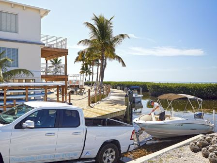 A white pickup truck is parked by a waterfront house, with a small boat on a trailer ready to launch into the calm water.