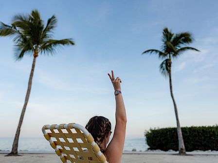 A person relaxing on a beach chair with two palm trees, blue sky, and ocean in the background, making a peace sign with one arm raised.