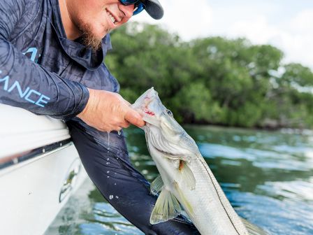 A man on a boat shows off a caught fish, smiling while the water below sparkles in the sunny scene.