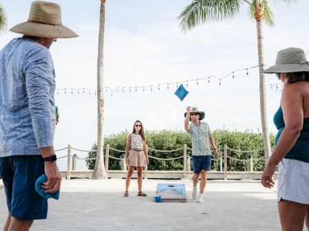 Friends play corn hole on a sunny beach with palm trees, as a wooden platform awaits the toss.