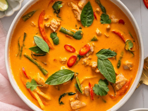 Creamy orange curry soup with chicken, bell peppers, chilis, and fresh basil in a white bowl, served with lime wedges and rice nearby.