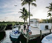 A sunny marina scene with palm trees, boats docked at a wooden pier, people chatting by the water, and a calm canal in the background.