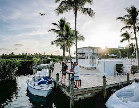 A sunny marina scene with palm trees, boats docked at a wooden pier, people chatting by the water, and a calm canal in the background.