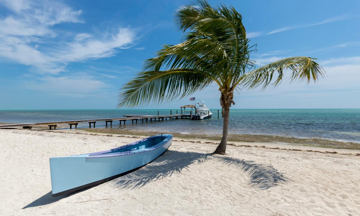 A sunny beach with white sand, a blue boat ashore, a leaning palm tree, and a wooden pier extending into calm turquoise water.